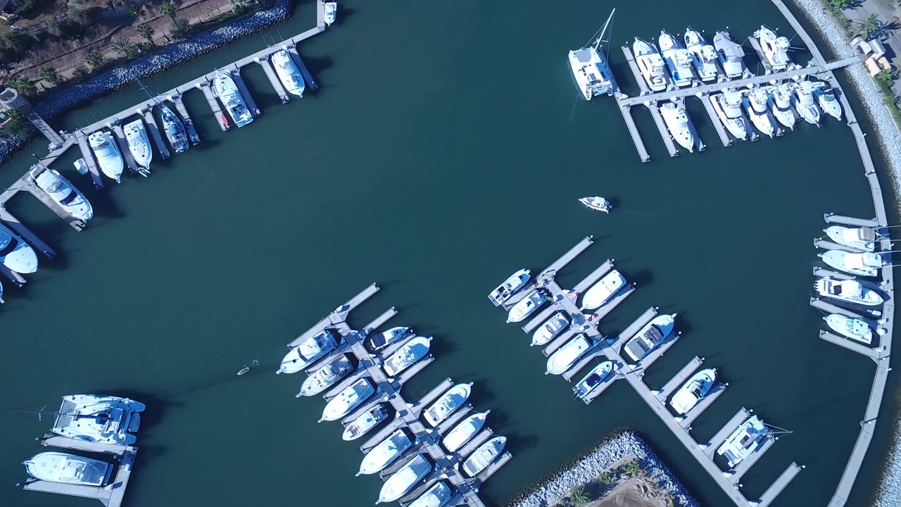 Aerial top-down view in Playita, Baja California Sur with clusters of white boats around marina slips and a small open patch of blue-green water, no visible people.