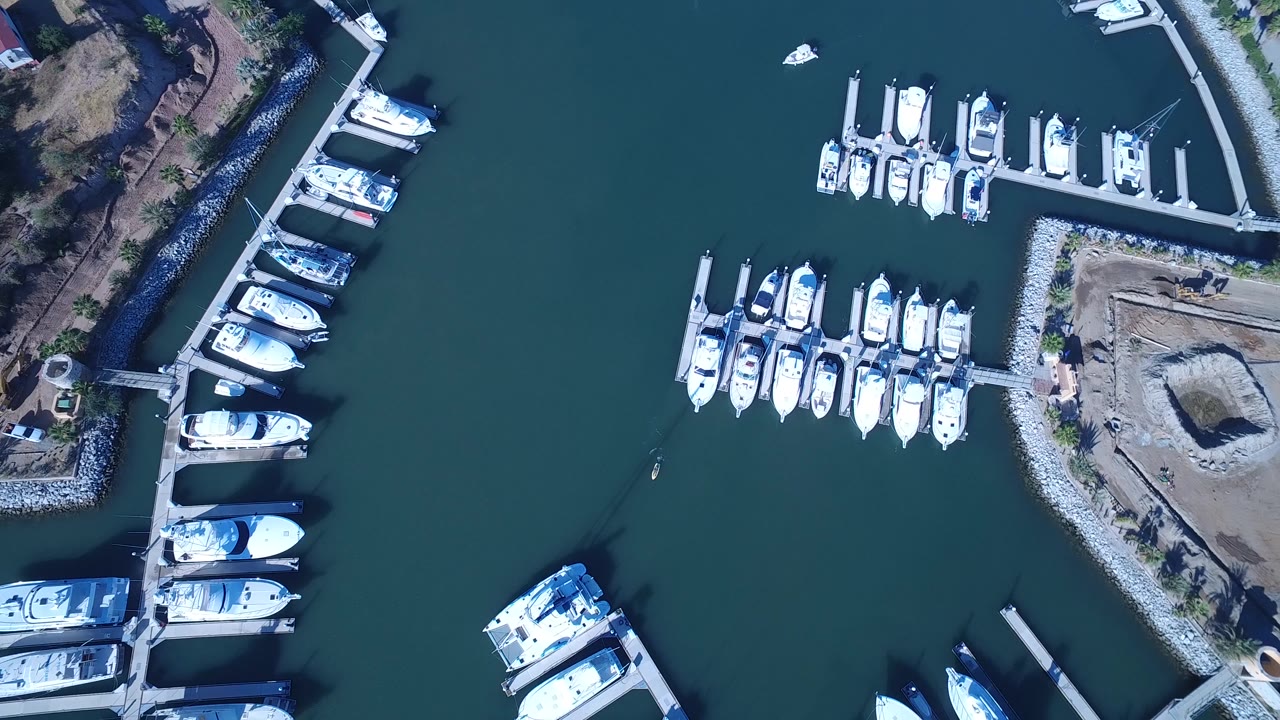 Aerial frame over Playita, Baja California Sur showing rows of white yachts in slips, a curved rock breakwater edge, and a bare lot beside the docks with tiny people visible.