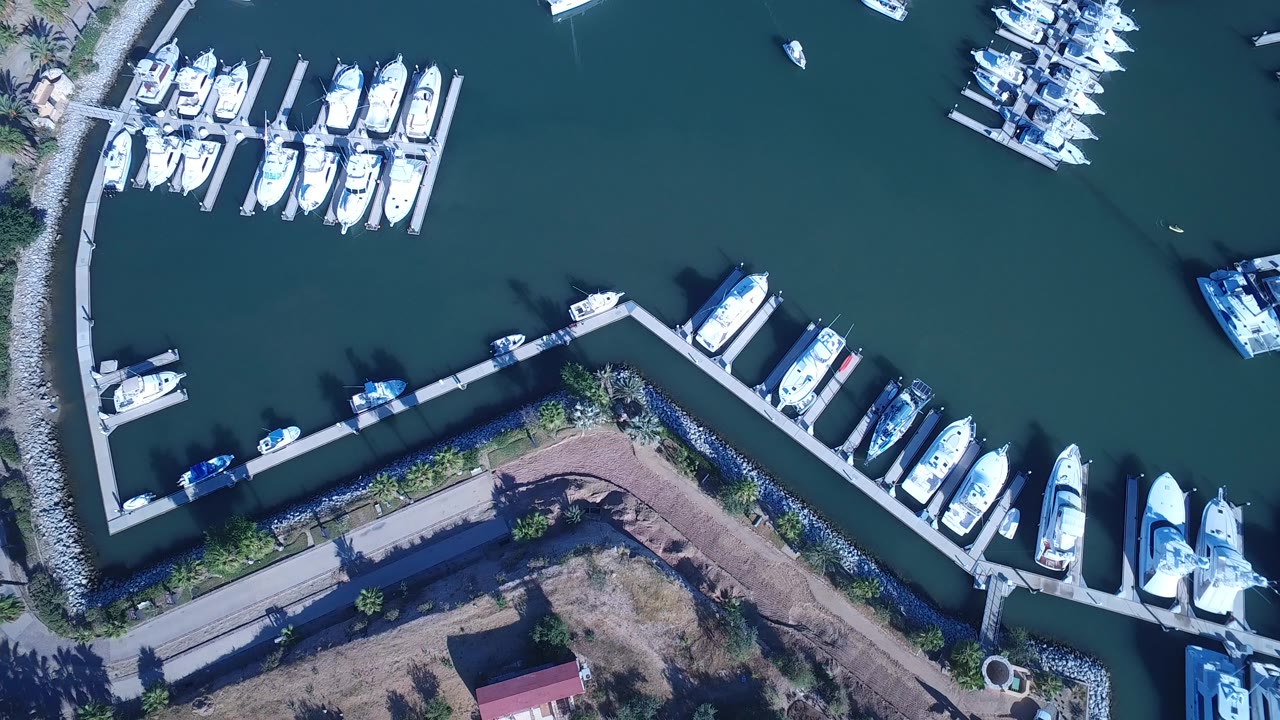 Overhead aerial frame of Playita, Baja California Sur, with white boats moored along angled docks, teal harbor water, a rock breakwater, shoreline road, and no visible people.