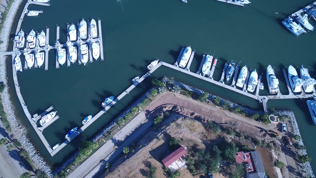Overhead frame in Playita, Baja California Sur showing white boats clustered along two dock sections, open teal water above, a curved stone breakwater at left, dry shoreline below, and no visible people.