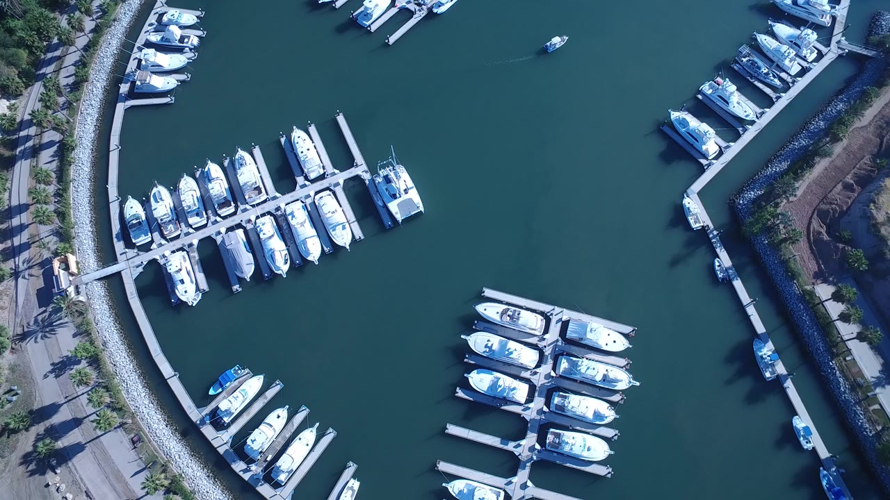 Overhead frame of Playita, Baja California Sur marina showing white boats around docks, a curved rock breakwater, green-blue water, shoreline paths, and no visible people.