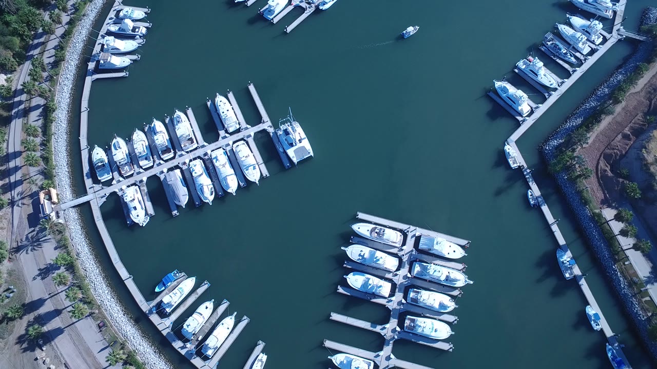 Top-down Playita marina view with the curved breakwater running along the left side, clustered white boats in slips, open green-blue water, and no visible people.