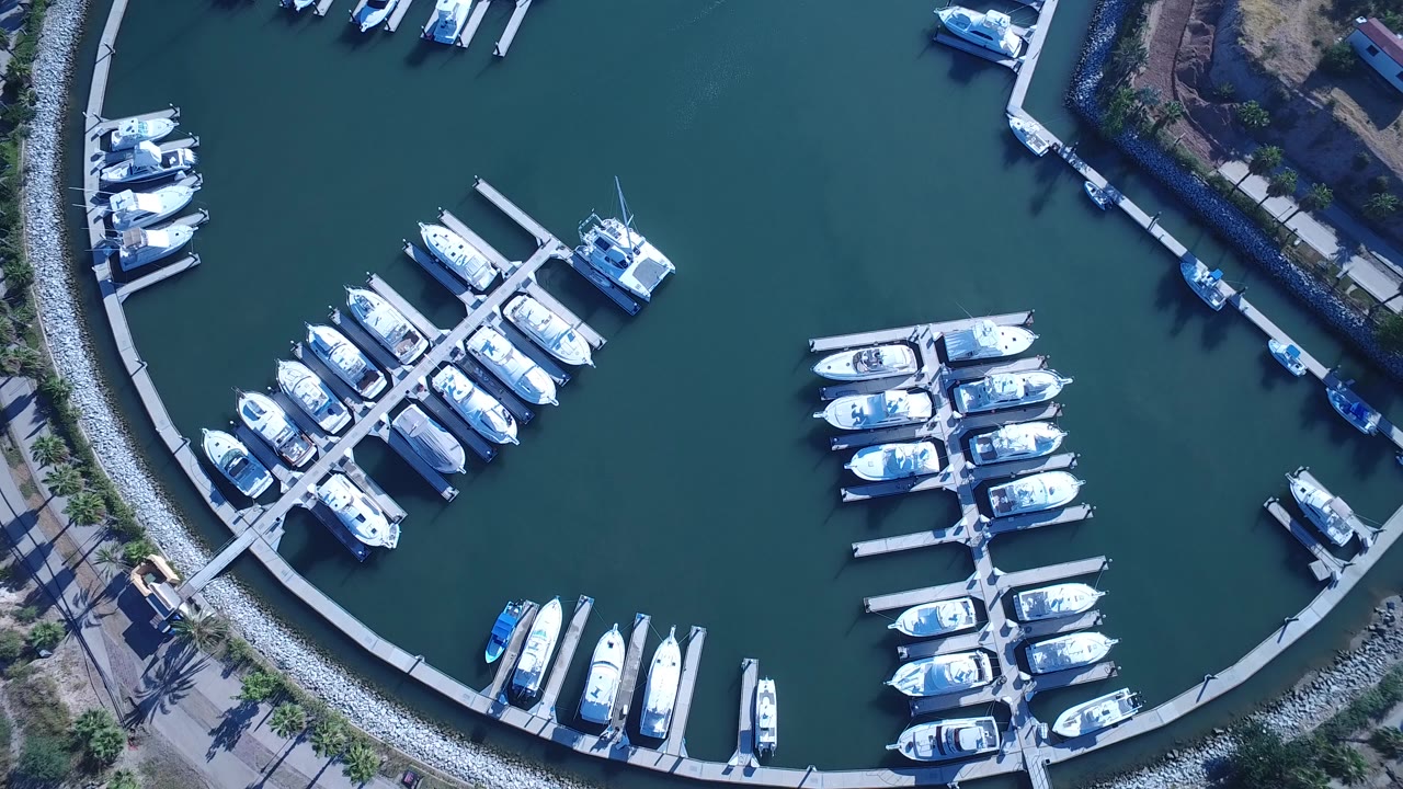 Top-down aerial view of docked white boats in Playita, Baja California Sur, with curved marina edges, green-blue water, and no visible people.