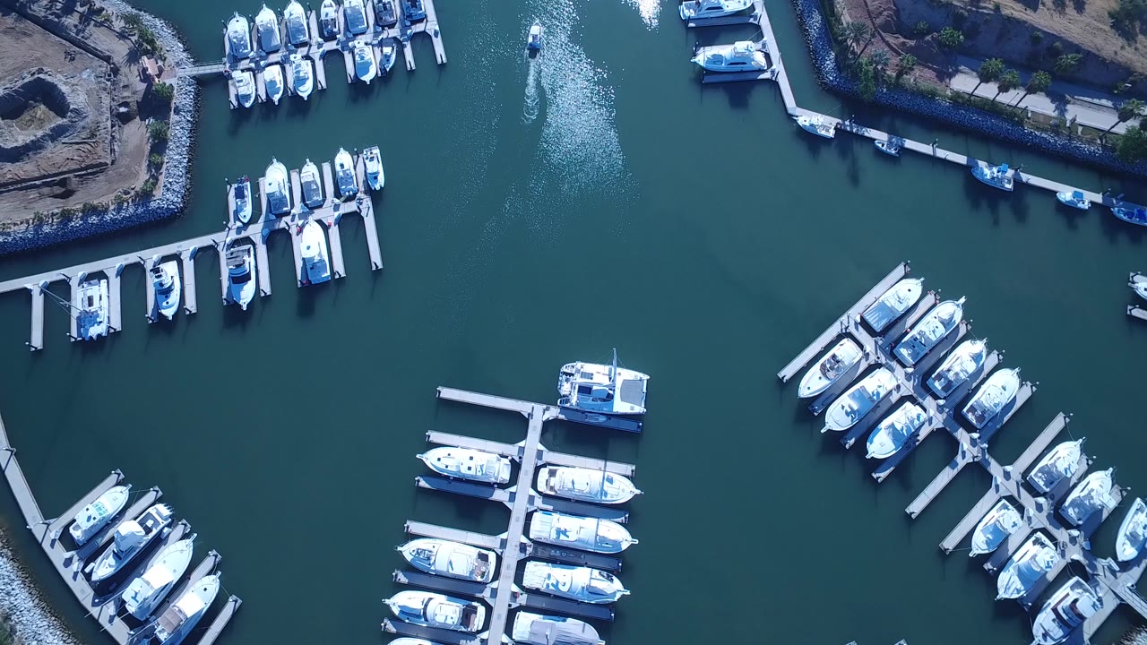 Overhead drone frame of a Playita, Baja California Sur marina with white boats docked in slips, teal water, rocky shoreline edges, and no visible people.