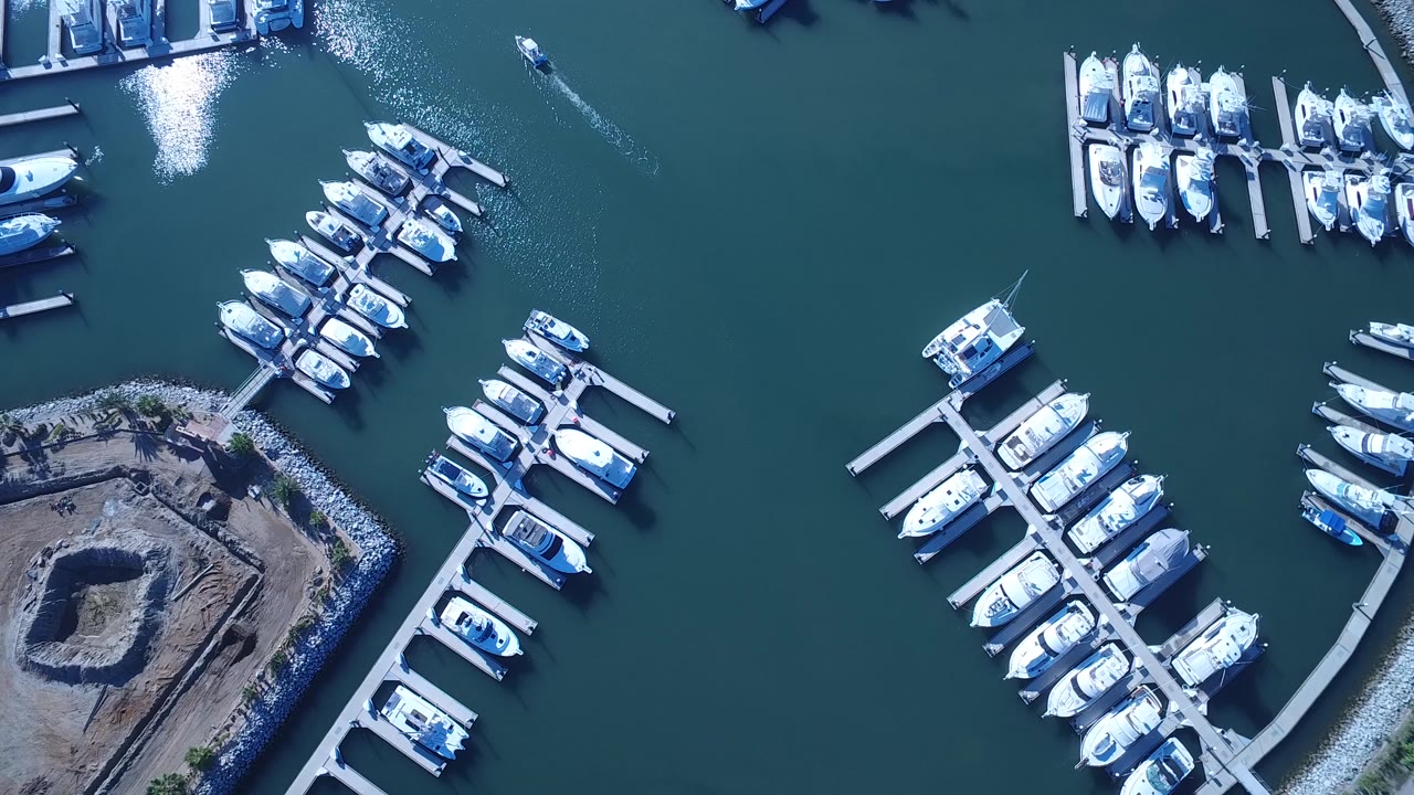 Aerial frame over Playita showing clustered white boats, angled docks, a rocky breakwater, dark teal water, and no visible people.