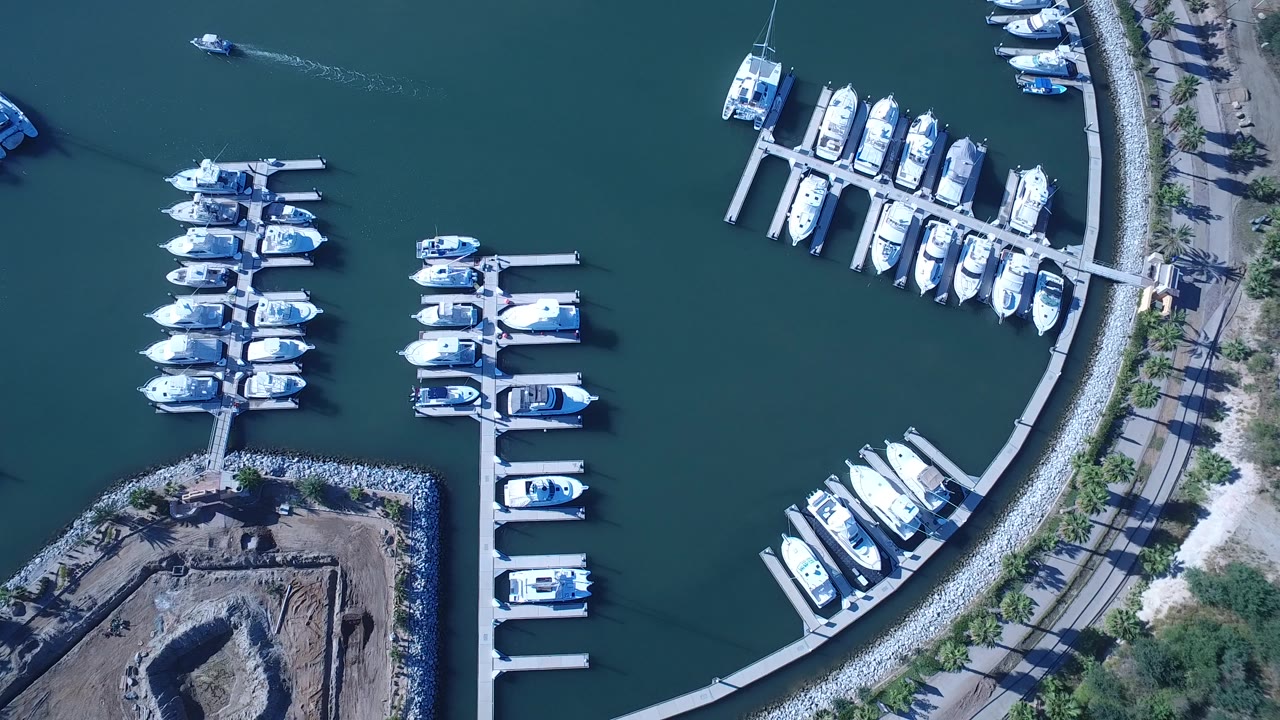 Aerial frame over a Playita marina showing rows of docked white boats, open blue-green water, a small boat near the upper left leaving a wake, and no visible people.