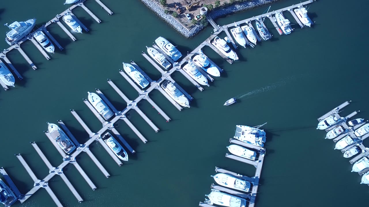High aerial frame over Playita marina showing docked white boats, empty slips, and a small boat moving across dark teal water with a thin wake behind it.