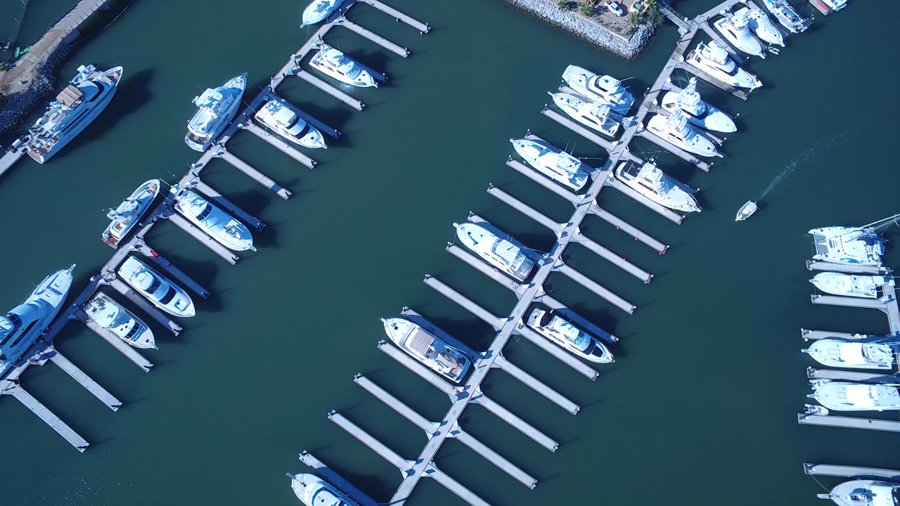 Overhead view of Playita, Baja California Sur marina docks with white boats moored in rows, dark teal water, a shoreline edge at left, and no visible people.
