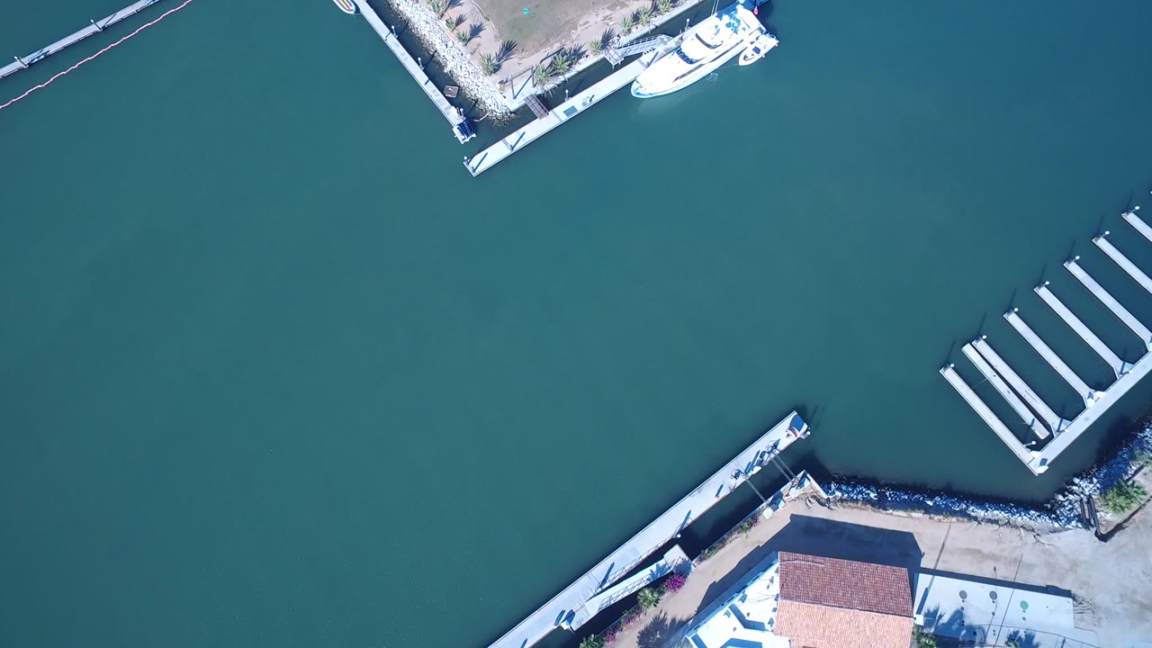 Overhead aerial frame from Playita, Baja California Sur showing green-blue water, partial marina docks, seawall corners, and no visible people.
