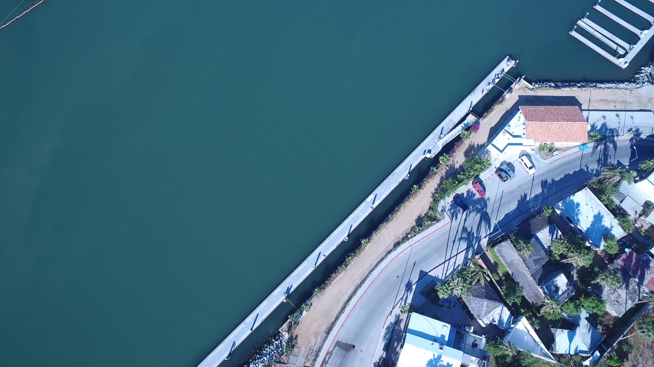 At 0:01, the drone angle stays high over Playita with the seawall running diagonally between broad teal water and a small cluster of homes, with no visible people.