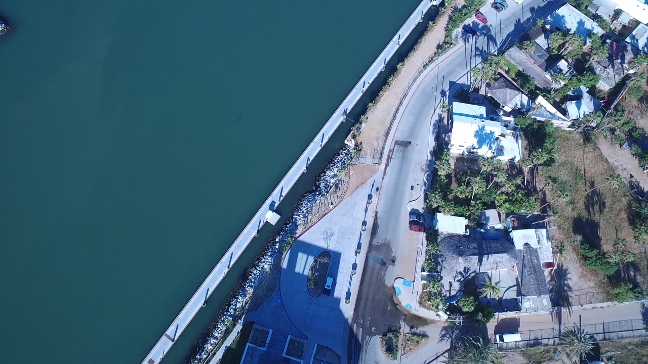 High overhead frame in Playita, Baja California Sur, Mexico, with the bridge or seawall on the left, a small dark car on the road, shoreline buildings, palm shadows, and no visible people.