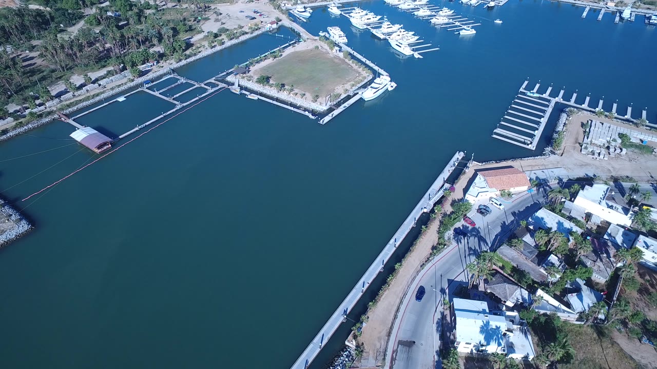 Aerial frame over Playita, Baja California Sur, showing blue water, a curved shoreline road, white marina docks, floating pens, low buildings, palms, and no visible people.