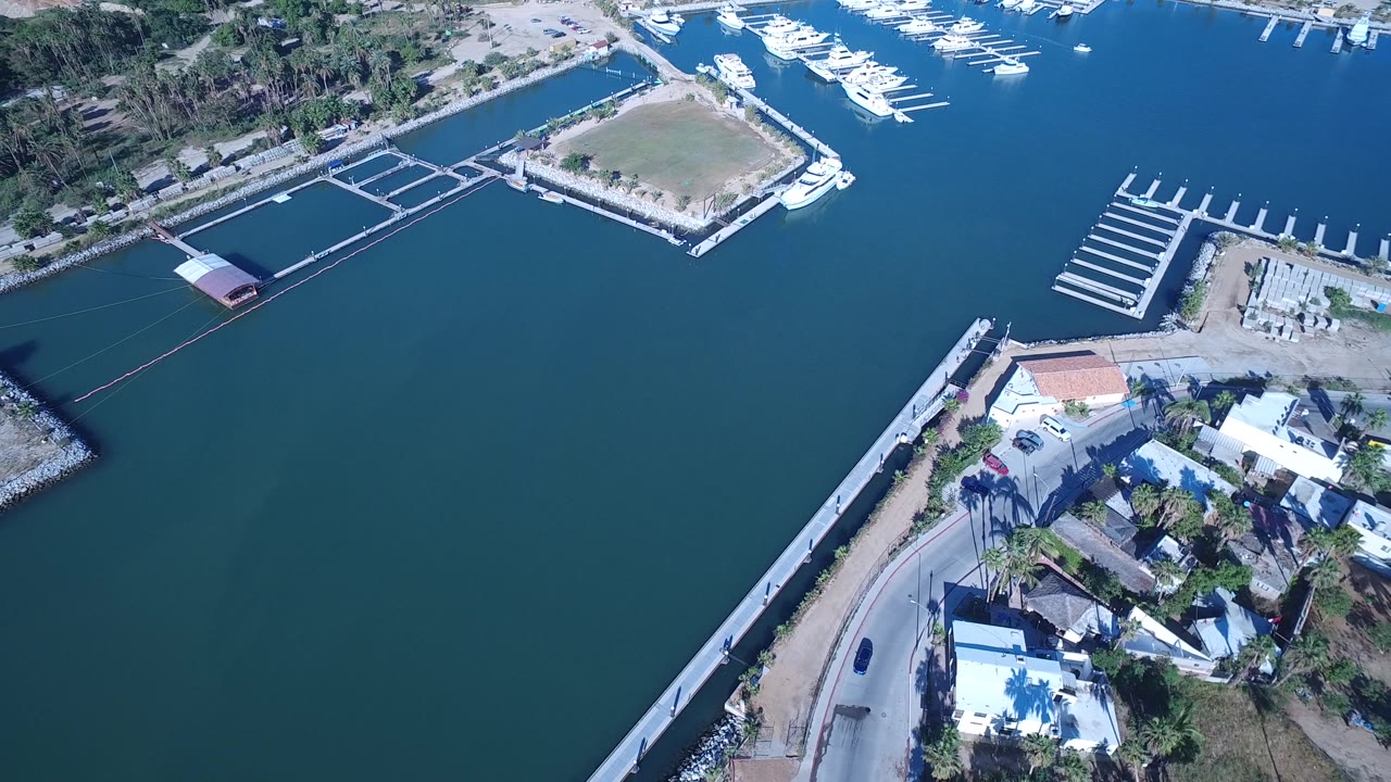 Aerial view across blue water toward docks, boats, and a road along the shore.