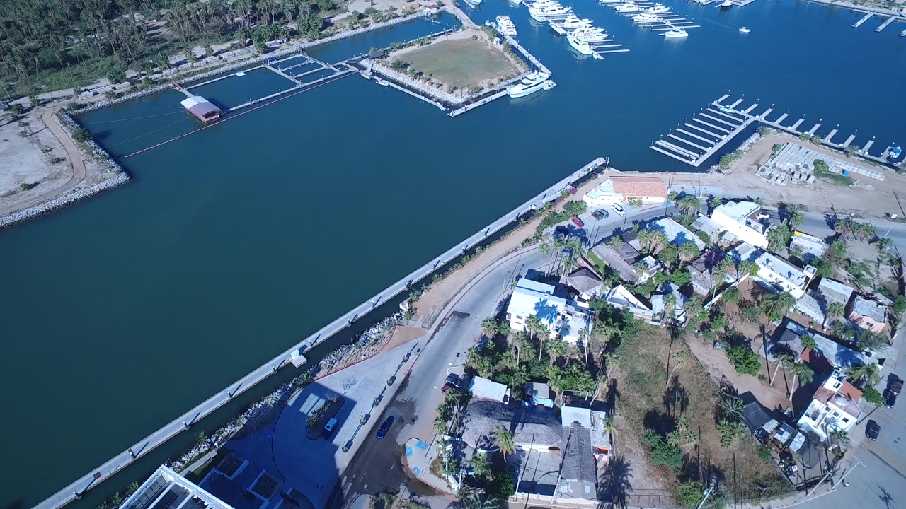 Aerial frame in Playita, Baja California Sur, with the canal filling the upper left, low buildings and palms in the midground, docks at the far side, and no visible people.