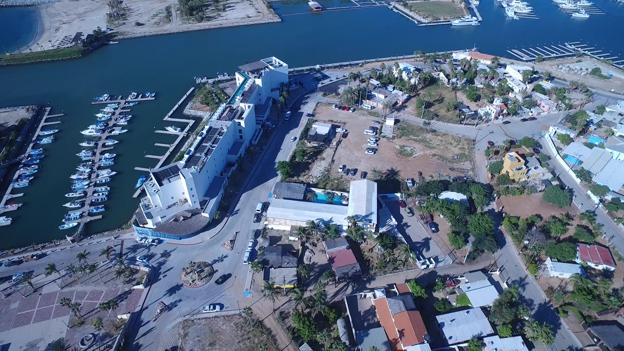 High aerial view over Playita, Baja California Sur, Mexico, showing a white waterfront hotel-like building, marina slips with docked boats, roads, parked cars, and nearby homes, with no visible people.