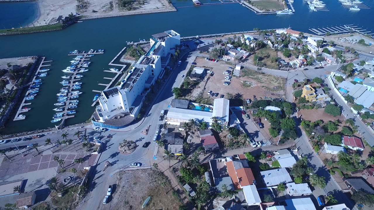Oblique aerial of marina water, a waterfront hotel-like structure, boats, homes, and roads.