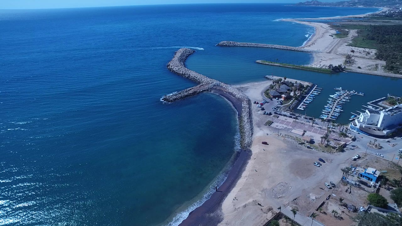 Opening aerial frame over San Jose del Cabo, Mexico, with blue ocean, a curved rock breakwater, sandy beach, marina docks, white coastal buildings, and no visible people.