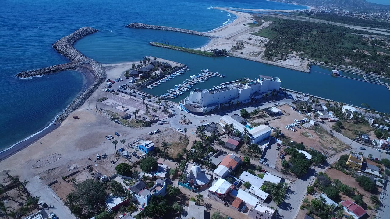 Aerial view of a coastal neighborhood beside a marina channel, beach, ocean, and rock breakwaters.