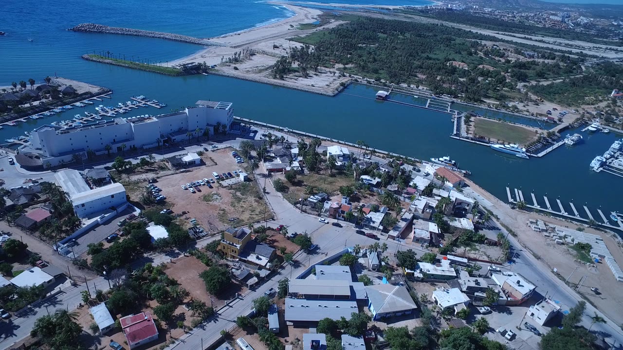 Aerial frame in San Jose del Cabo showing more of the marina channel, clustered homes in the lower frame, waterfront buildings at left, and no visible people.