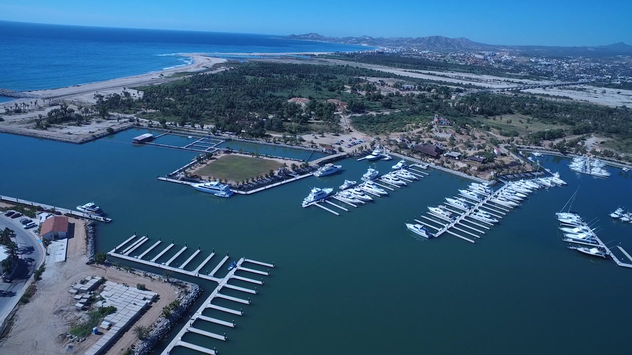 Second frame in San Jose del Cabo, Mexico holds above the marina with empty finger piers in the lower foreground, docked white boats at center, beach and ocean beyond, and no visible people.