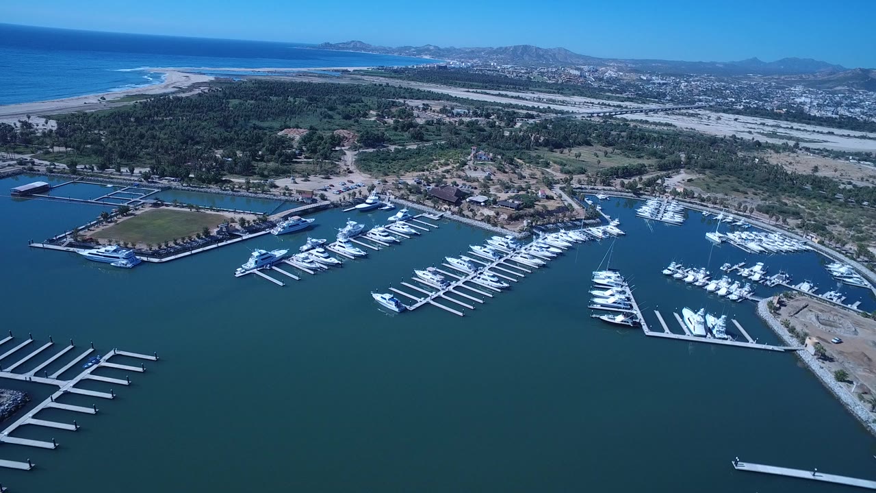 Opening aerial frame over a San Jose del Cabo marina with white boats, empty docks, dark water, beach, ocean, town, hills, and no visible people.
