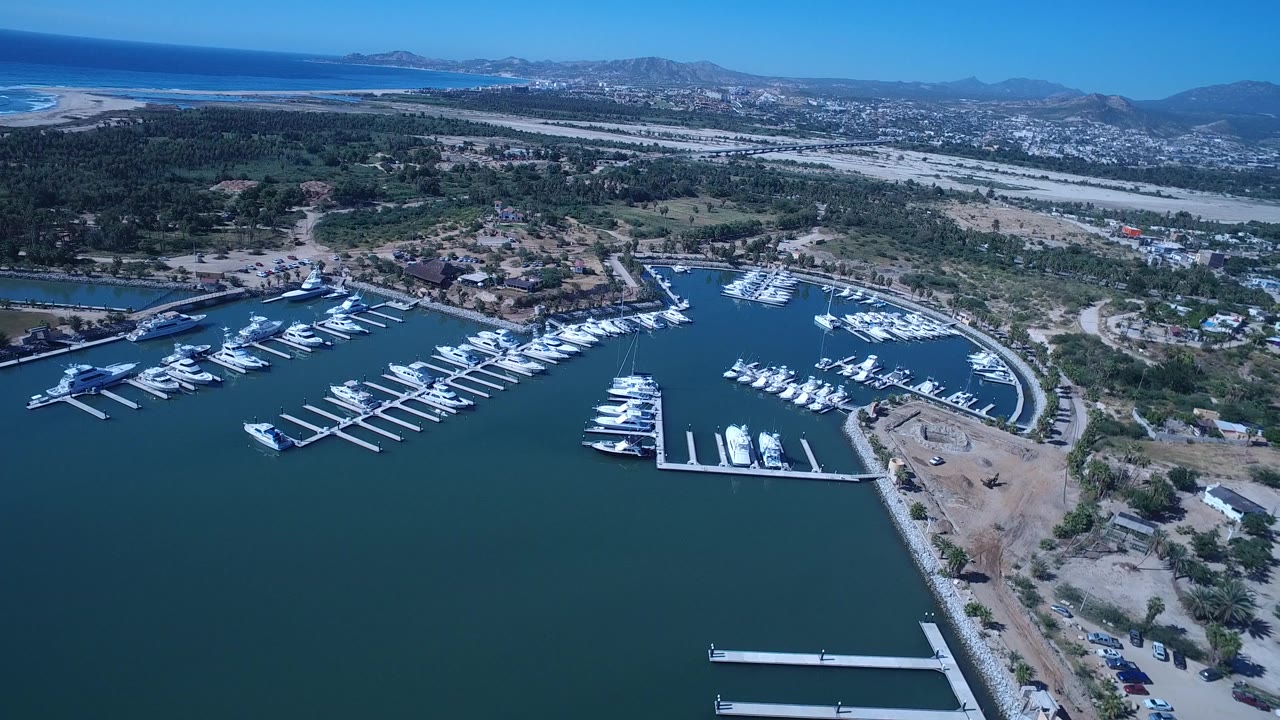 A San Jose del Cabo marina fills the lower frame with rows of white boats, curved shore roads, dry terrain, ocean surf, mountains, and no visible people.