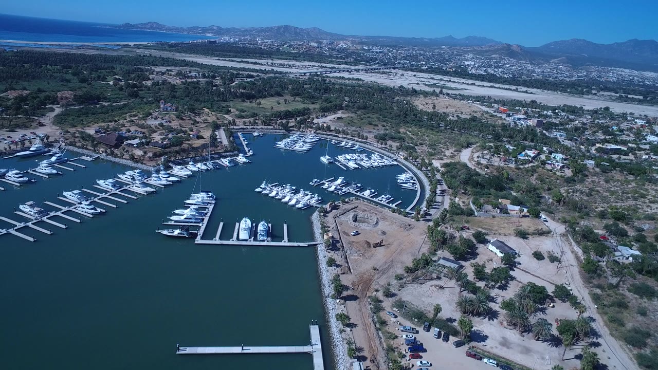 Aerial frame over San Jose del Cabo showing white boats at marina docks, dark water in the foreground, scattered houses and dry scrubland beyond, mountains on the horizon, and no clearly identifiable people.