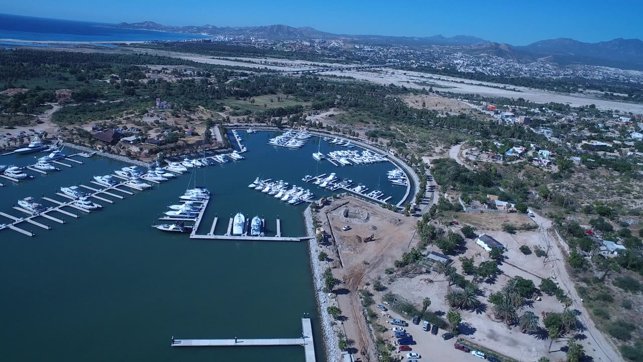 Aerial view in San Jose del Cabo with a curved marina basin, rows of docked boats, dry shoreline lots, inland houses, pale flats, distant mountains, and no clearly identifiable people.