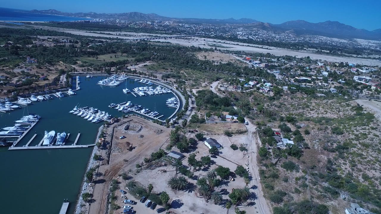 Aerial frame from San Jose del Cabo, Mexico, with the marina basin and docked boats on the left, scrub-covered ground and roads across the middle, hills on the horizon, and no visible people.