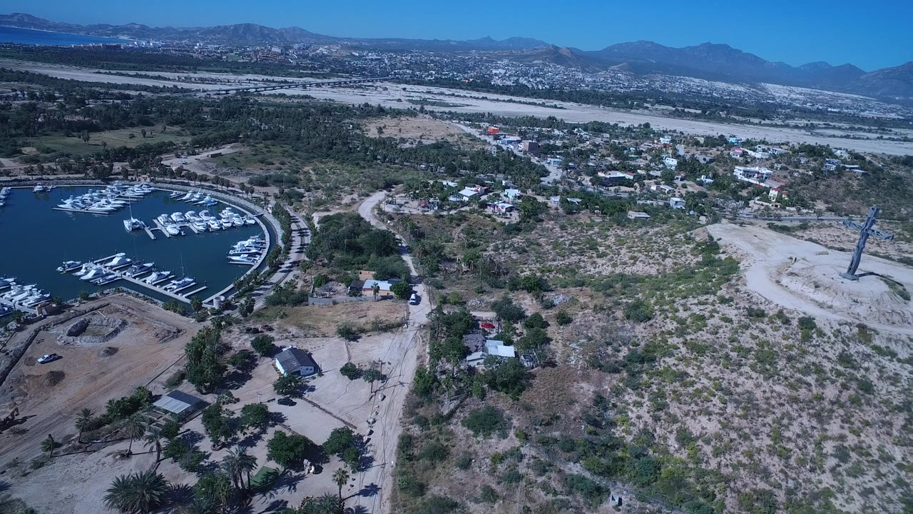 At 0:01, the San Jose del Cabo aerial holds the marina at the left edge while brushy open lots, dirt roads, small houses, pale flats, and distant mountains fill the frame with no visible people.