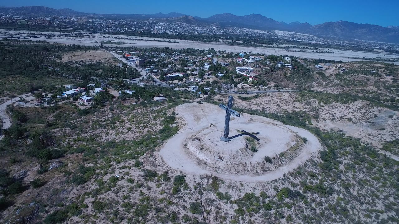 Wide aerial frame over San Jose del Cabo, Mexico, showing a large cross on a pale hilltop with scrubland, nearby buildings, distant mountains, and no visible people.