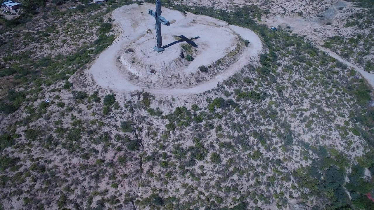 Aerial frame in San Jose del Cabo, Mexico with the blue cross near the upper center and mottled green brush spreading across the dry slope below, with no visible people.