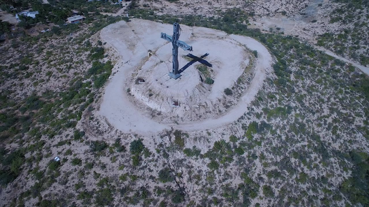 The San Jose del Cabo aerial frame keeps the cross centered on the pale circular hill, with green brush and dry terrain extending beyond it and no visible people.