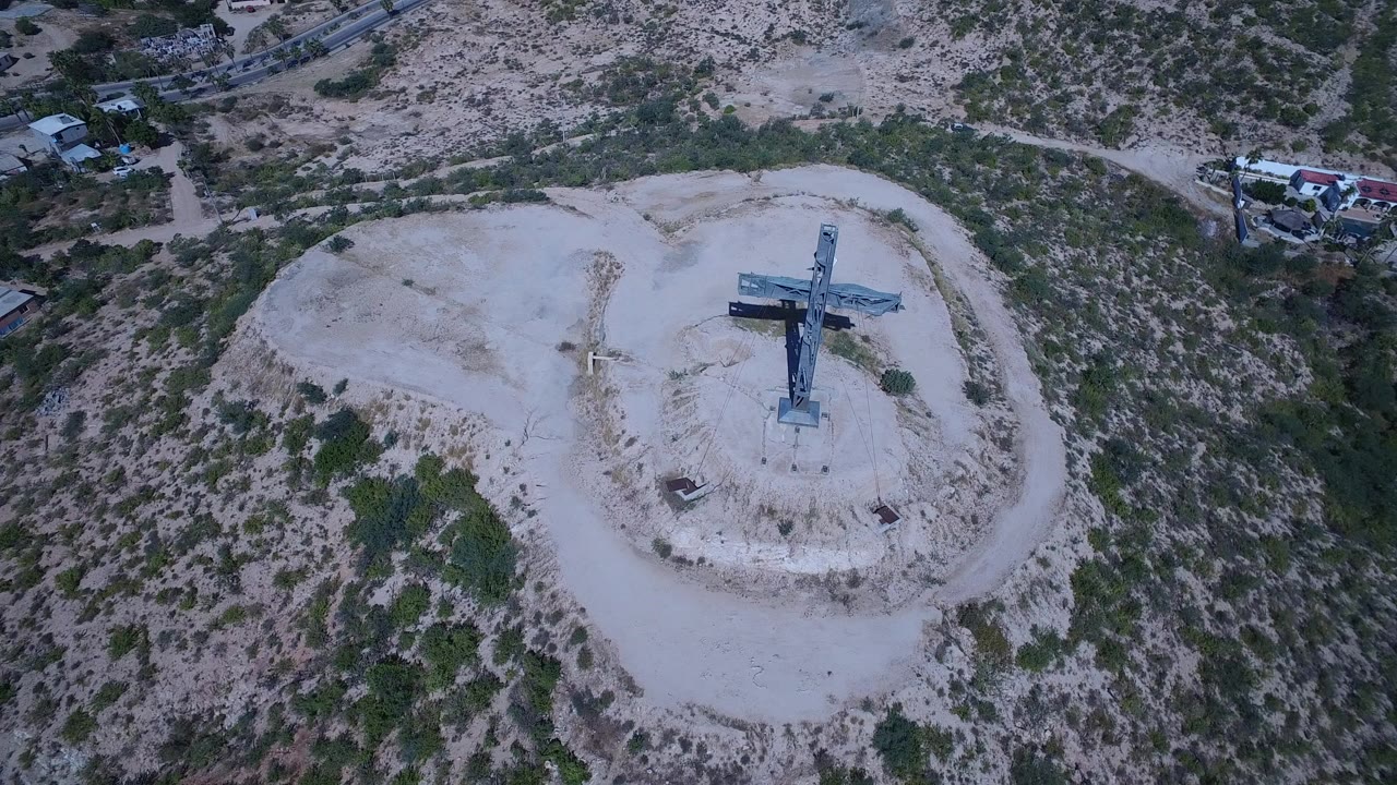 Aerial frame from San Jose del Cabo, Mexico, showing a blue cross centered on a pale hilltop with scrub vegetation around it and no visible people.