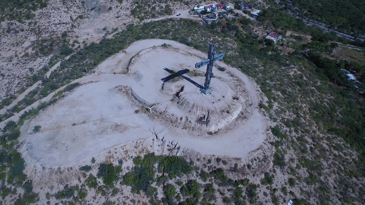 Aerial view of a hilltop cross monument on pale dirt surrounded by scrub vegetation.
