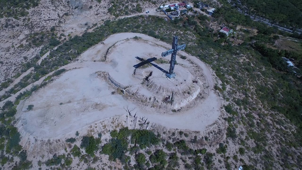 Aerial frame in San Jose Del Cabo, Mexico, with the cross monument near the upper center of a rounded dirt mound, green brush on the slopes, distant structures, and no visible people.
