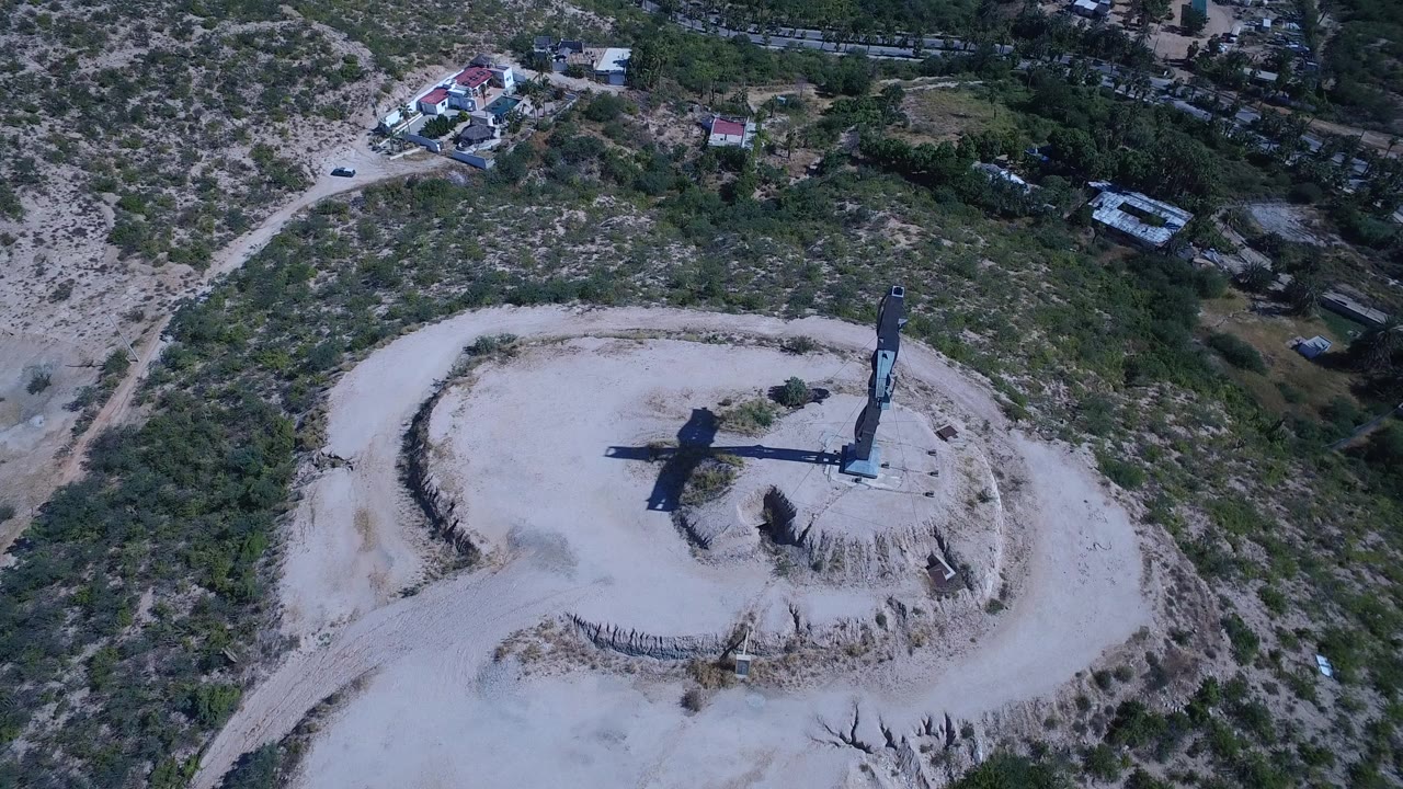 High aerial view in San Jose del Cabo, Mexico, showing a tall cross monument on a pale circular platform with scrubby green terrain, sandy tracks, nearby buildings, and no visible people.