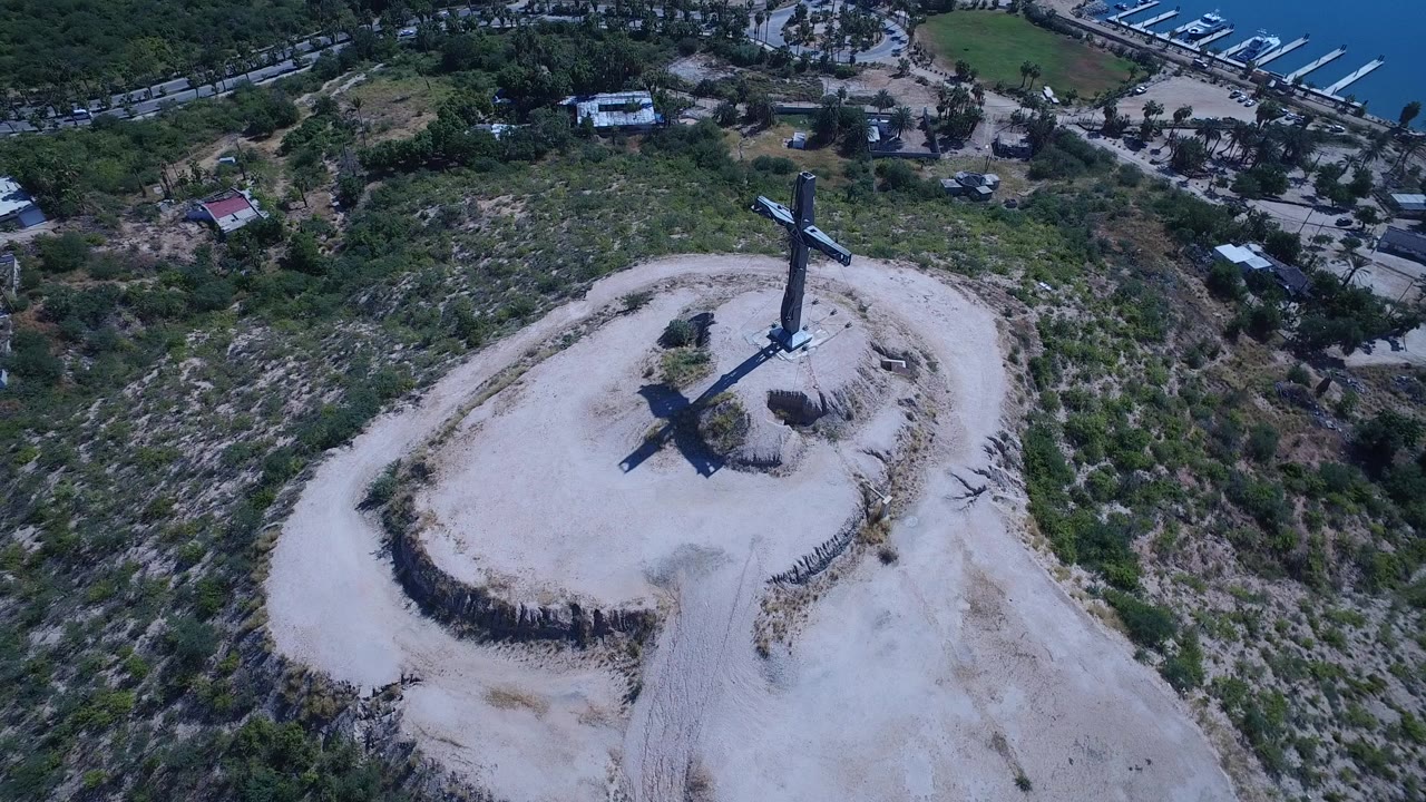 High aerial frame over San Jose del Cabo showing a large dark cross on a pale dirt hilltop, scrubby slopes, roads, blue marina water, and no visible people.