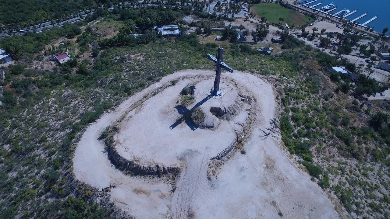 Aerial view in San Jose del Cabo with the cross centered on the dusty overlook, a curving access road around it, dry brush, marina slips, and no visible people.