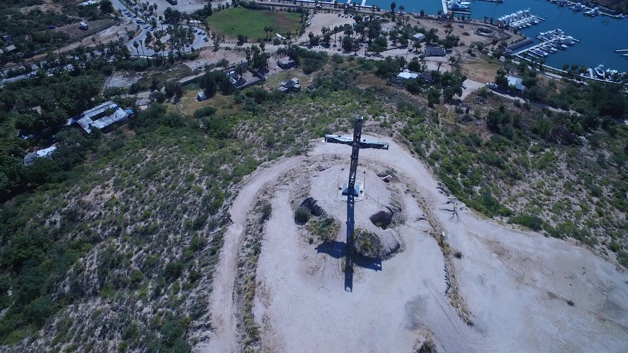 Aerial frame from San Jose del Cabo with the cross slightly lower in view, dusty ground and brush filling the hill, marina slips and blue water along the top edge, and no visible people.