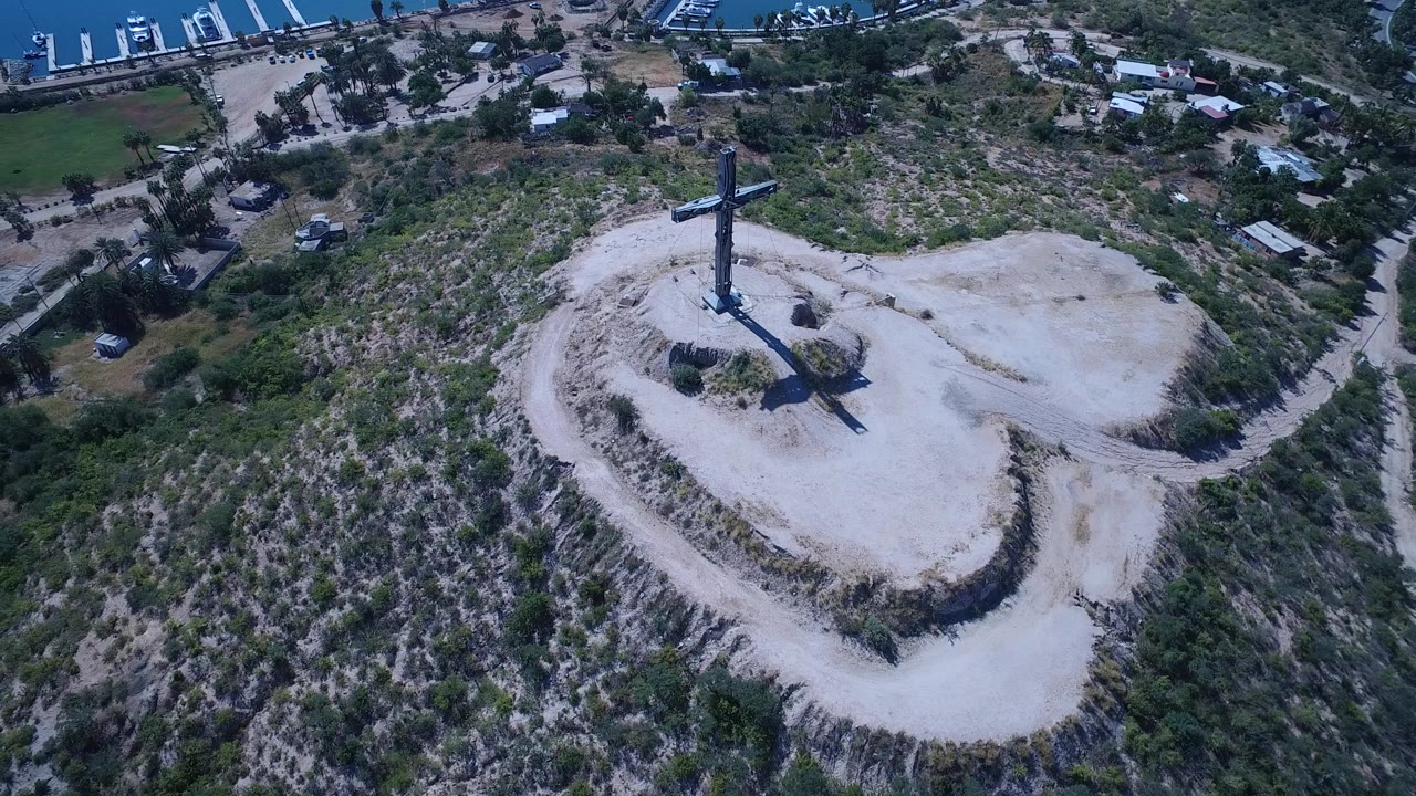 Aerial view of a tall cross on a pale dirt hilltop with scrubland around it