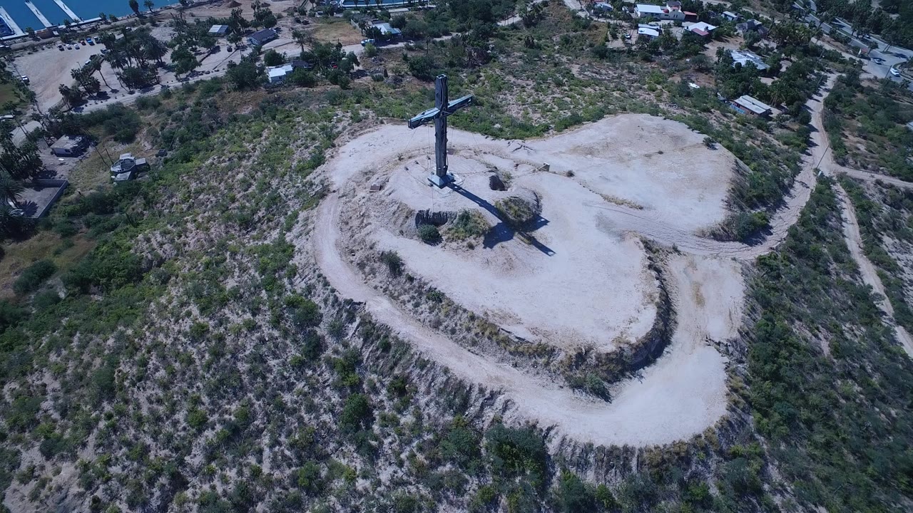 Drone-height view in San Jose del Cabo with the crucifixion monument slightly left of center, dry hillside paths below, small buildings beyond, and no visible people.