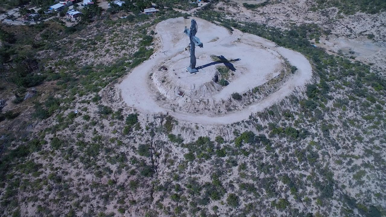 San Jose del Cabo aerial view with the cross near the top center, a curved dirt road around the mound, mottled green scrub below, and no visible people.