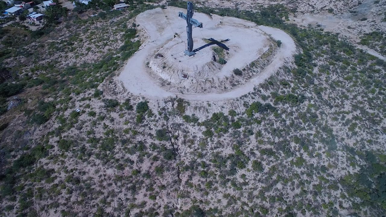 High aerial view in San Jose del Cabo with the crucifixion monument near the top of frame, dry scrubland below, and no visible people.