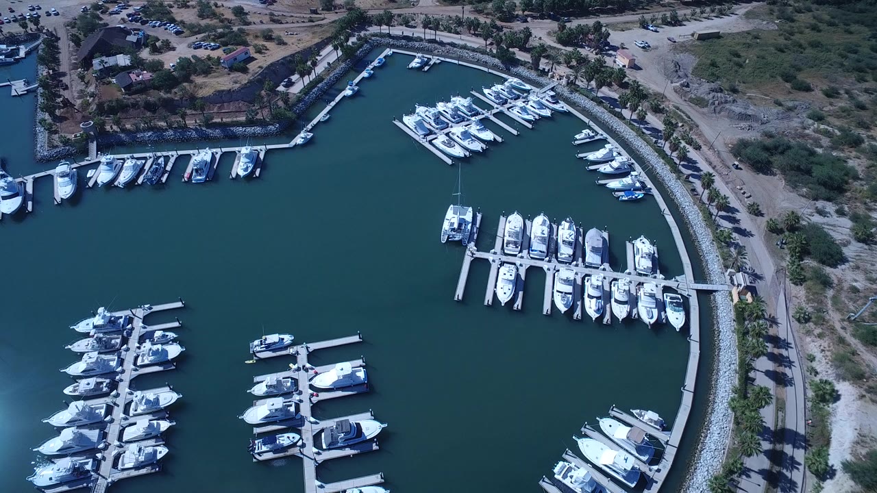 Overhead drone view of docked yachts and boats in a San Jose del Cabo marina, with blue-green water, curved shoreline, dry land, and no visible people.