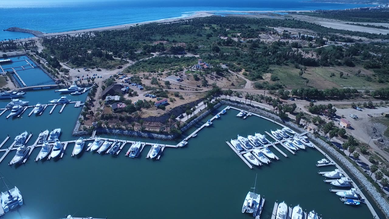 High aerial view over a San Jose del Cabo marina with white boats in slips, teal harbor water, roads, trees, beach, and ocean in the distance, with no visible people.