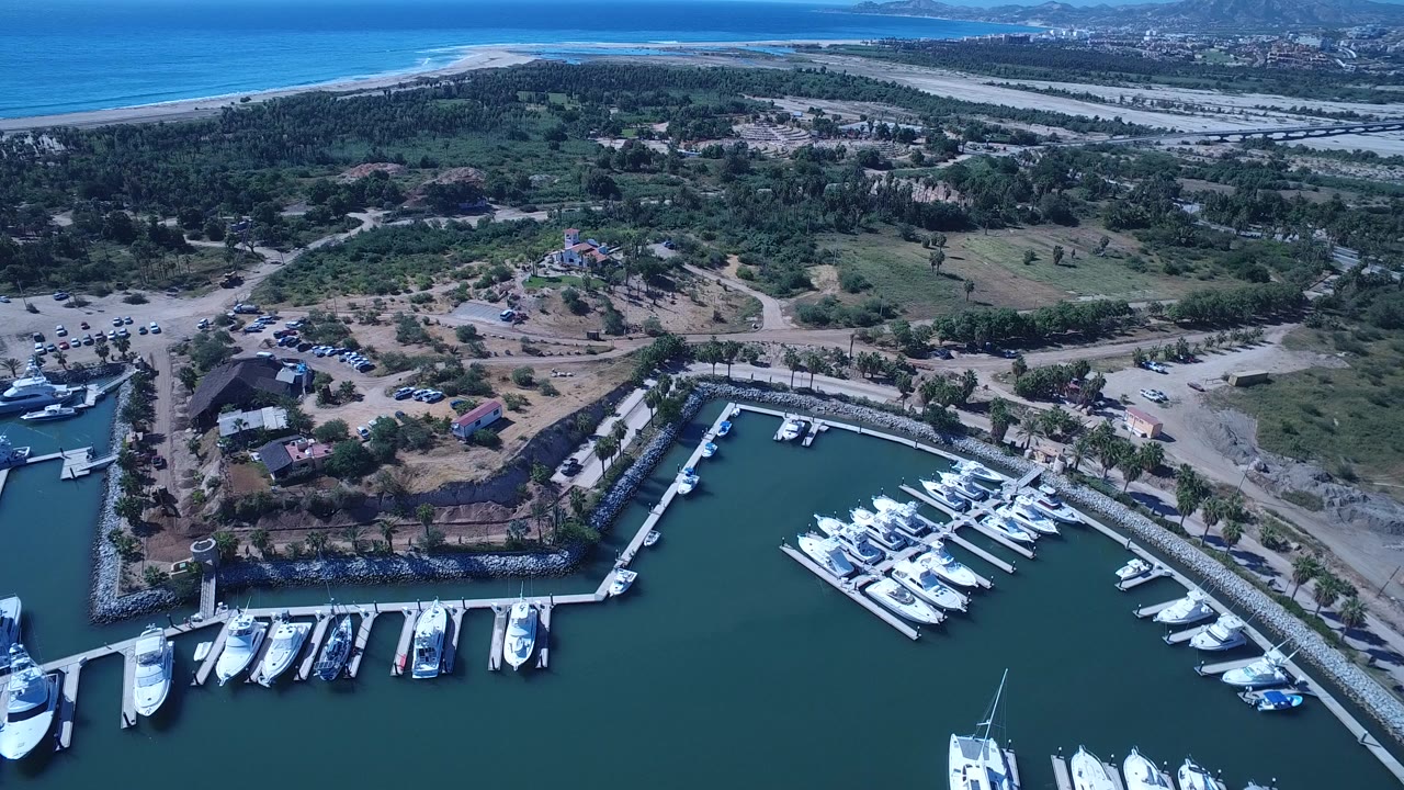 Aerial frame over San Jose Del Cabo, Mexico, showing white boats at marina docks in the foreground, scrubland and roads behind them, and beach with ocean beyond, with no recognizable people visible.