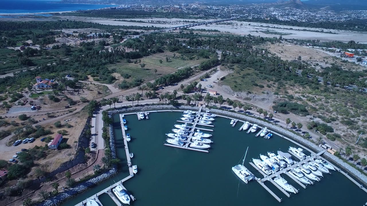 High drone view in San Jose del Cabo, Mexico, with the marina lower in frame, dry lots and curved roads behind it, scattered buildings in the distance, and no identifiable people.