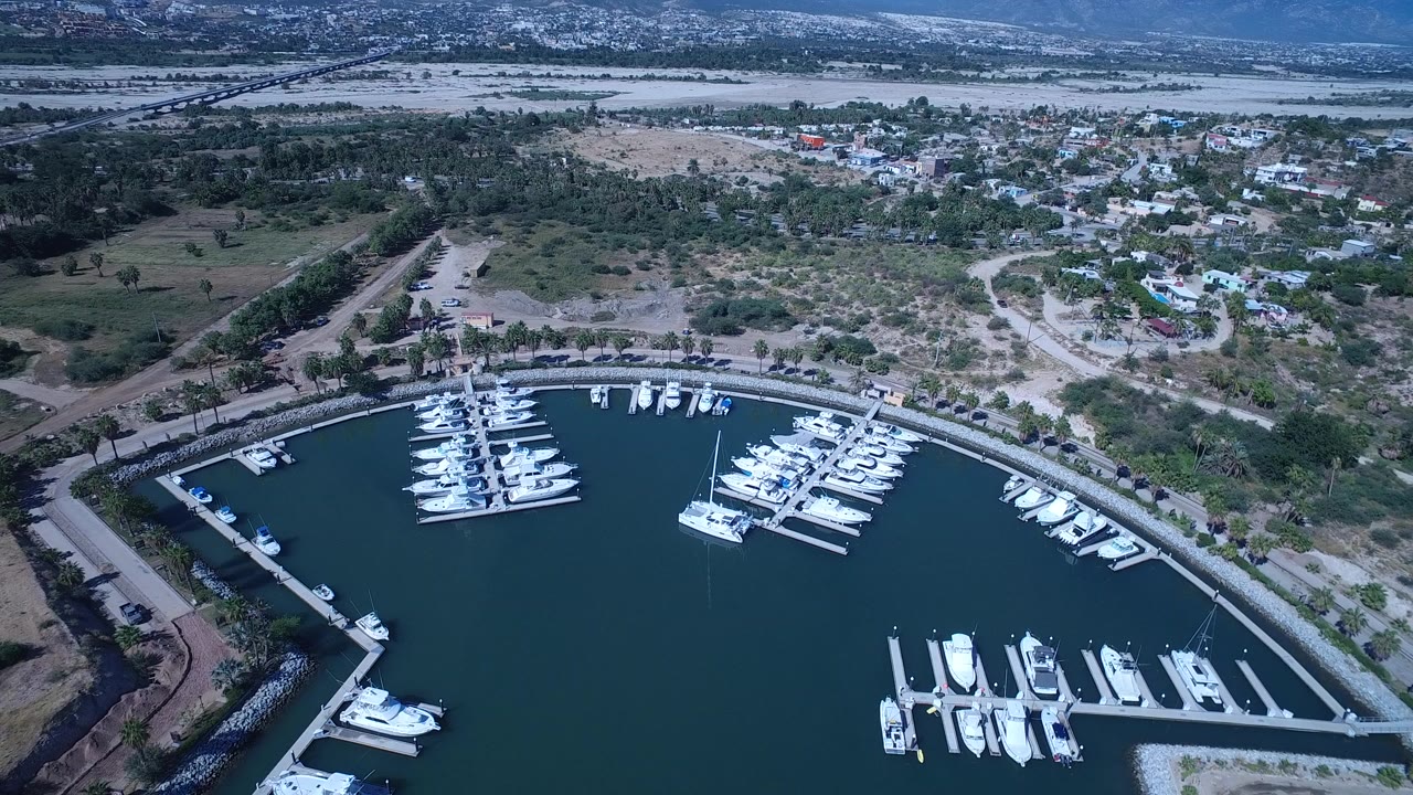 High aerial frame over a San Jose Del Cabo marina, with white yachts in dark water, curved docks, a shore road, dry open land, scattered homes, and no visible people.