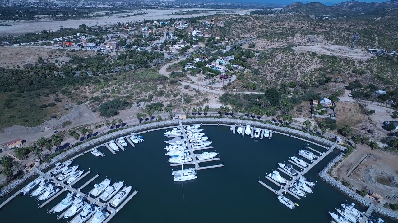 High aerial view over a San Jose del Cabo marina, with white boats in curved docks, dark water in the foreground, dry hills and scattered buildings beyond, and no visible people.